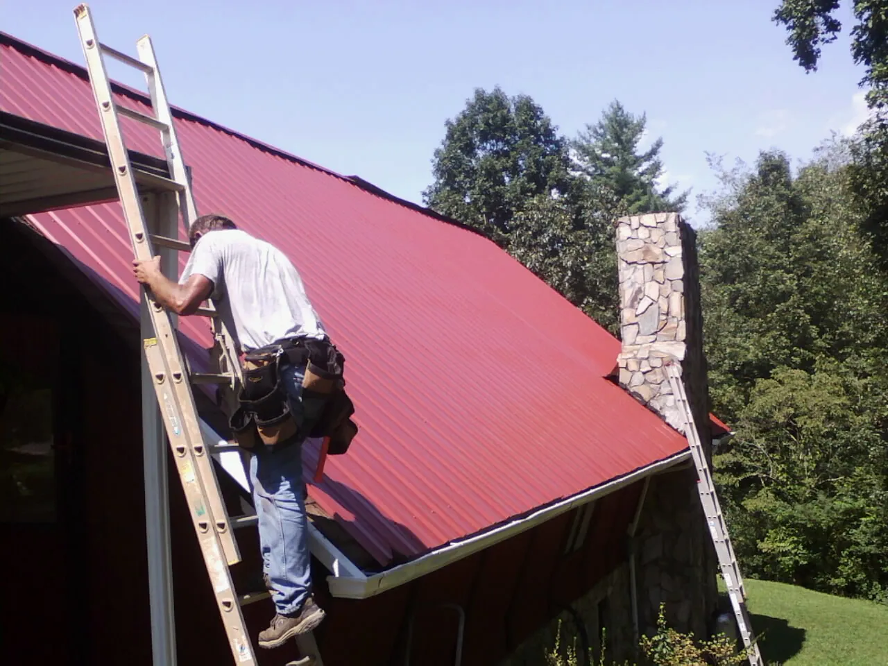 Roofer on a ladder beside a red metal roof and stone chimney — Soffit & Fascia Repair by Roofing Brothers in Decorah